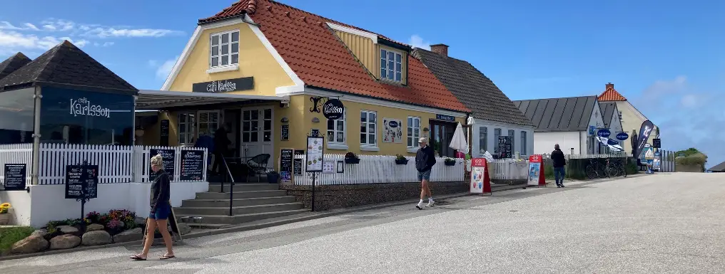 Café Karlsson – Kysten 1, Lønstrup Café Karlsson i Lønstrup – gul facade med rødt tag, hvid stakit og menutavler ved Kysten